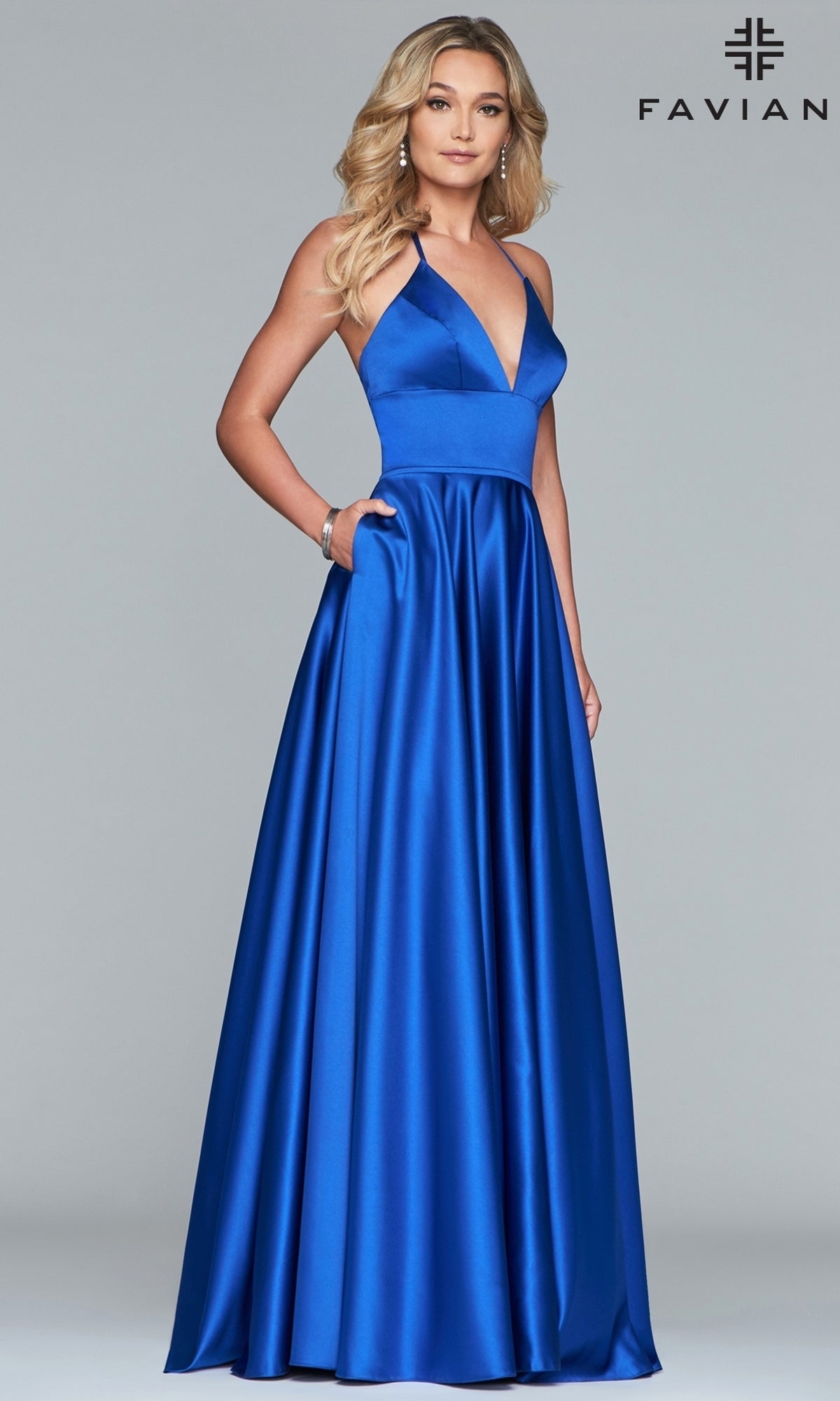 A woman wears the Corset-Back A-Line Long Satin Prom Dress by Faviana, a floor-length royal blue gown with a deep V-neckline and halter straps, standing against a plain gray background.